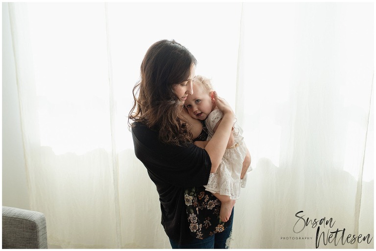 Mom stands near window and holds baby girl close as she rests her head on her mom